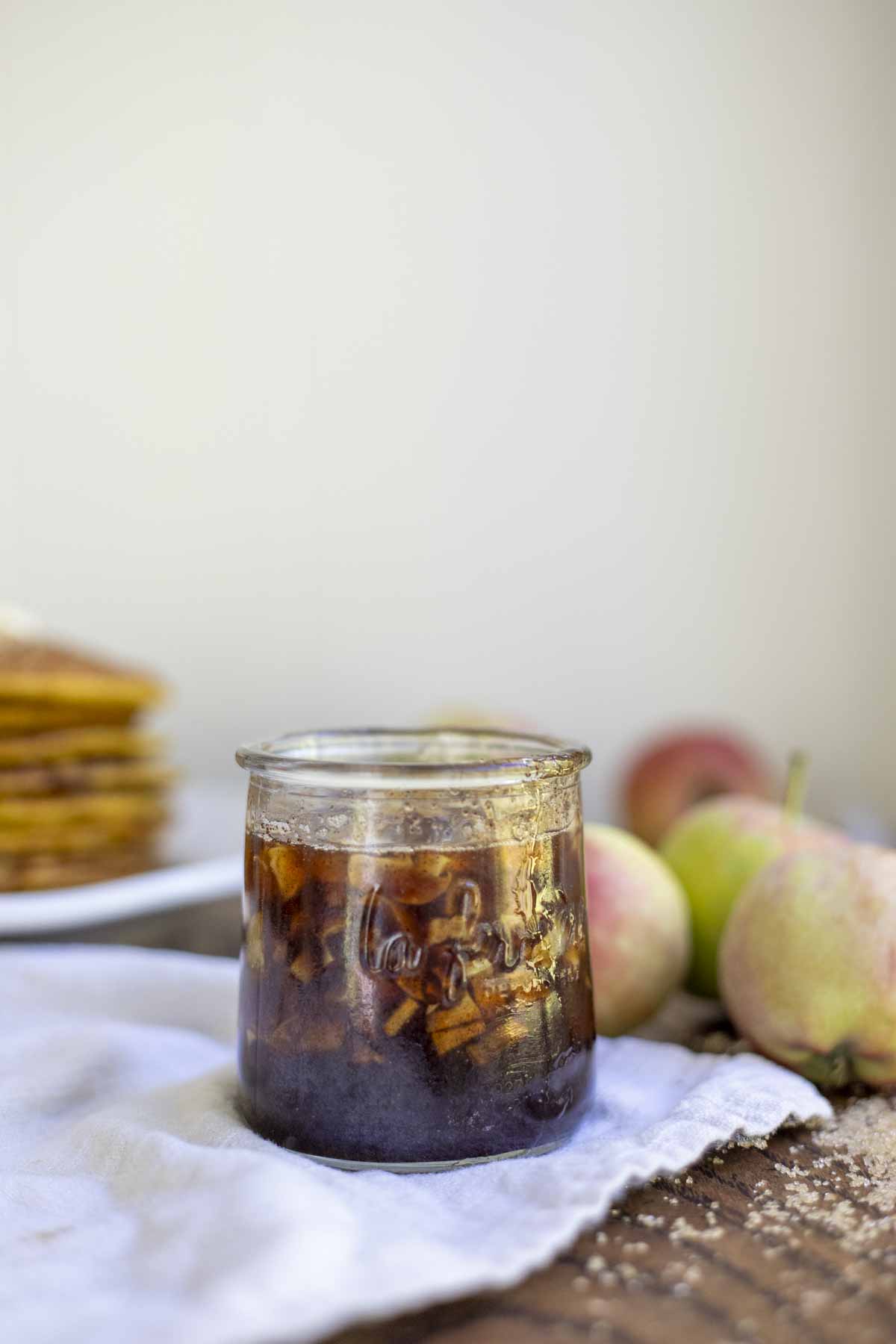 cinnamon apple syrup in a small glass jar on a napkin with apples and a stack of pumpkin pancakes in the background.