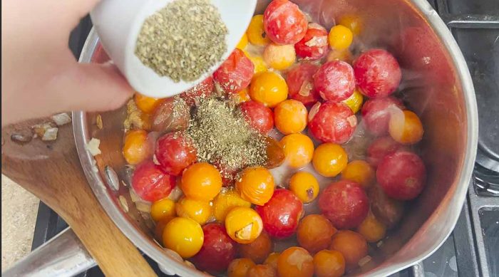 Italian seasoning being added to cherry tomatoes and onions in a pot
