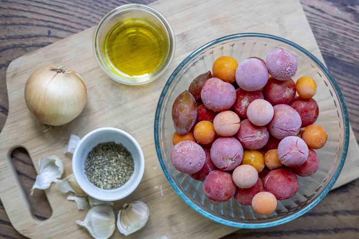 cutting board with onion, garlic, Italian seasonings in a white jar, olive oil in a glass jar and a bowl of frozen cherry tomatoes.