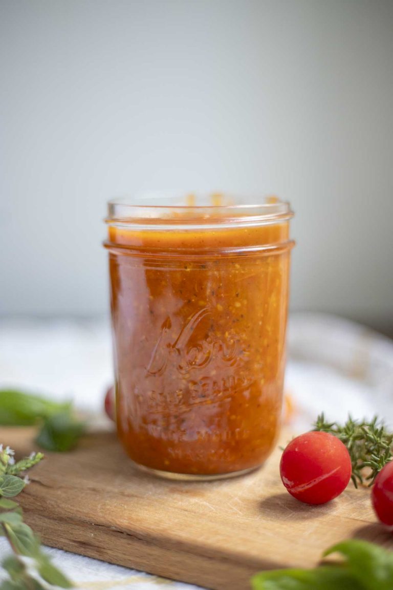 Cherry tomato sauce in a glass jar on a cutting board with fresh herbs and cherry tomatoes surrounding the jar