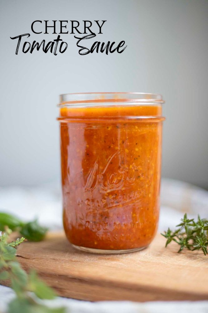 Cherry tomato sauce in a wide mouth mason jar on a wood countertop with herbs surrounding the jar