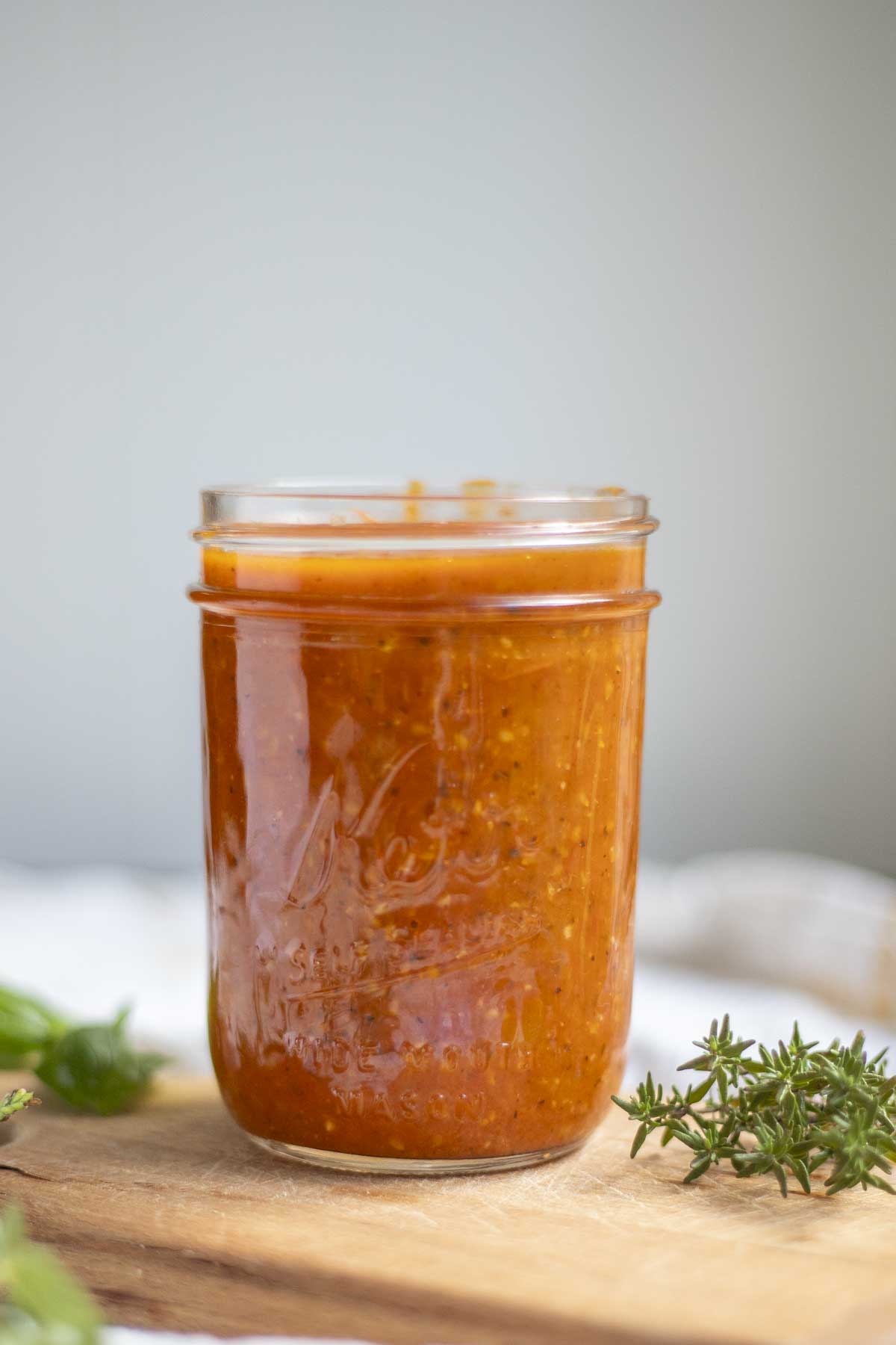 jar of cherry tomato spaghetti sauce on a wood cutting board surrounded by herbs.