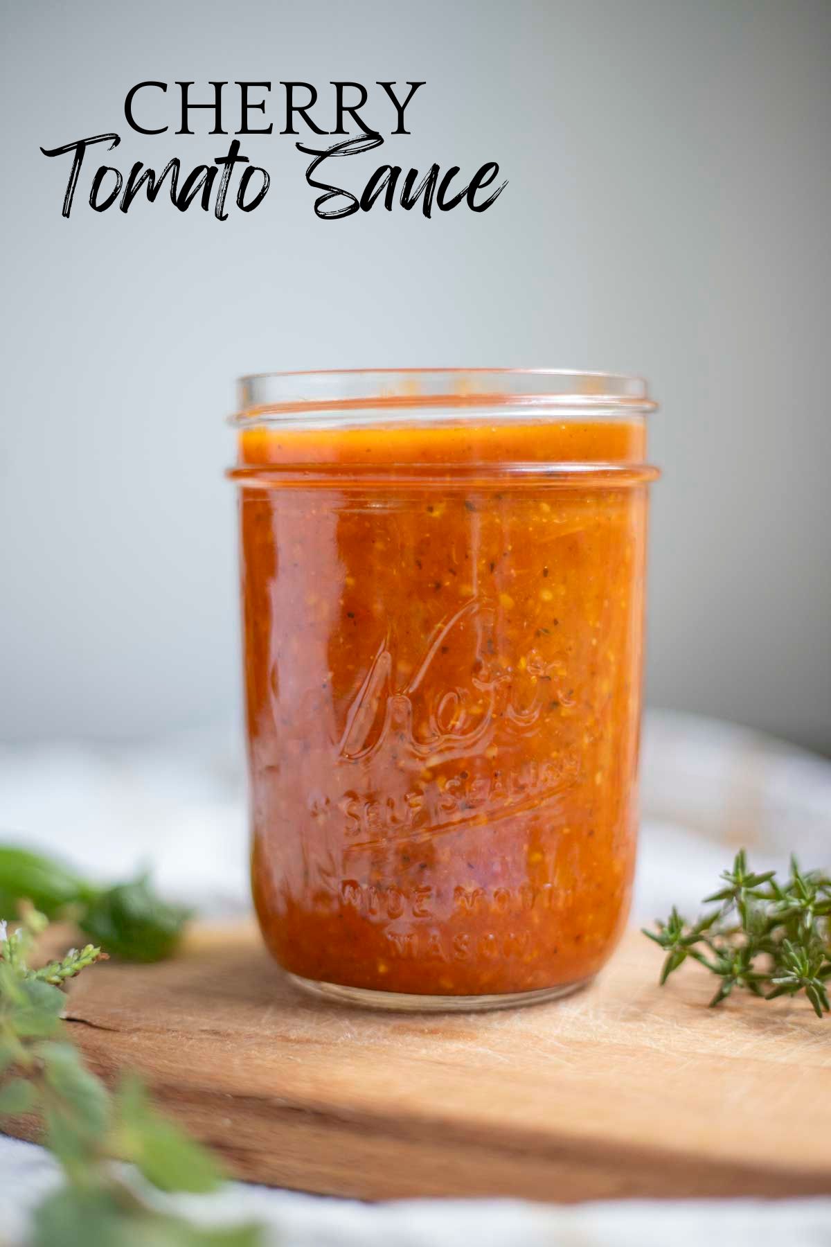 Cherry tomato sauce in a wide mouth mason jar on a wood countertop with herbs surrounding the jar.