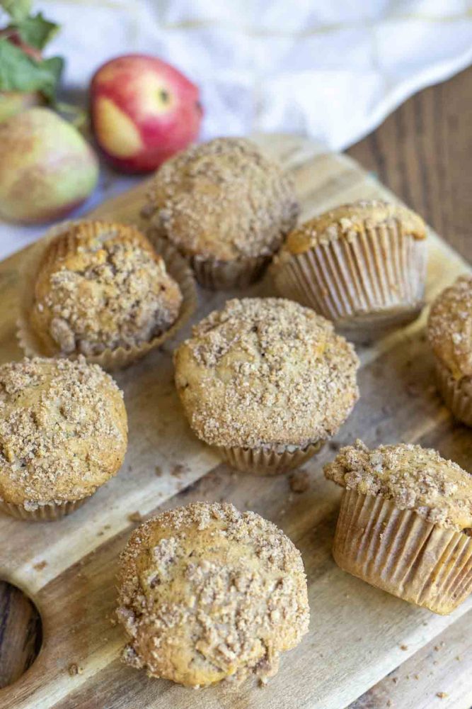 muffins scattered on a wood cutting board with apples and a white and yellow towel in the background