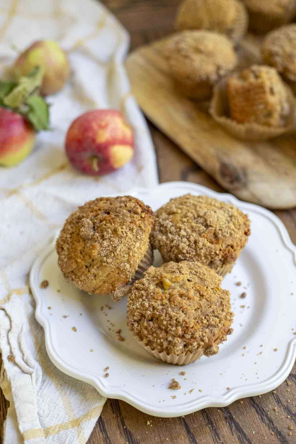 three sourdough apple muffins with a crumble topping on a white plate on a wood table with a yellow and white towel, apples, and more muffins on a cutting board in the background