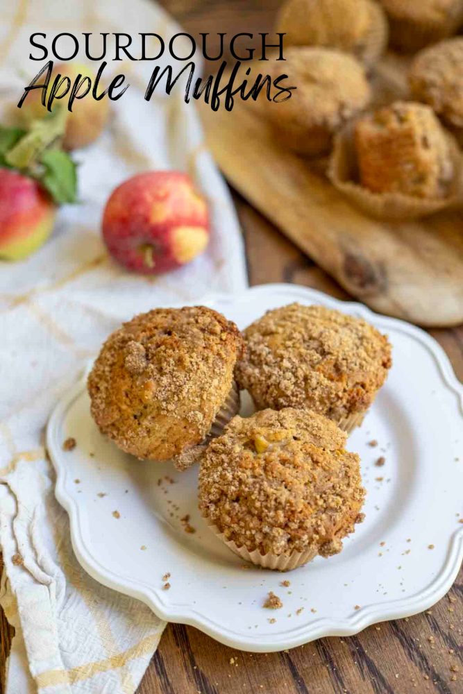 three sourdough apple muffins with a crumble topping on a white plate on a wood table with a yellow and white towel, apples, and more muffins on a cutting board in the background
