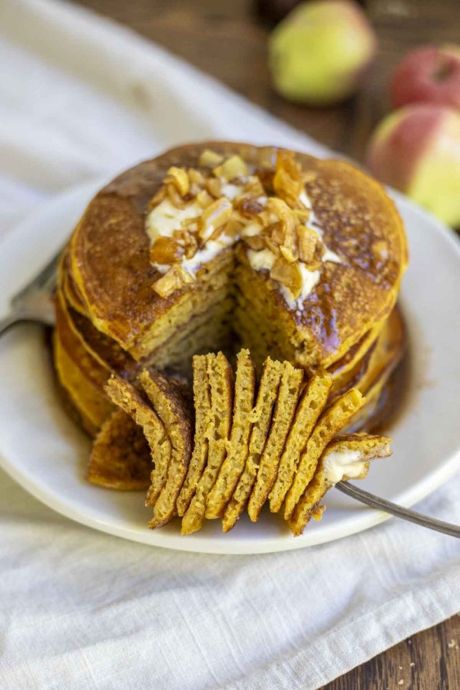 stack of sourdough pancakes topped with apples and syrup with a large slice of pancakes taken out and on a fork resting in front of the stack.