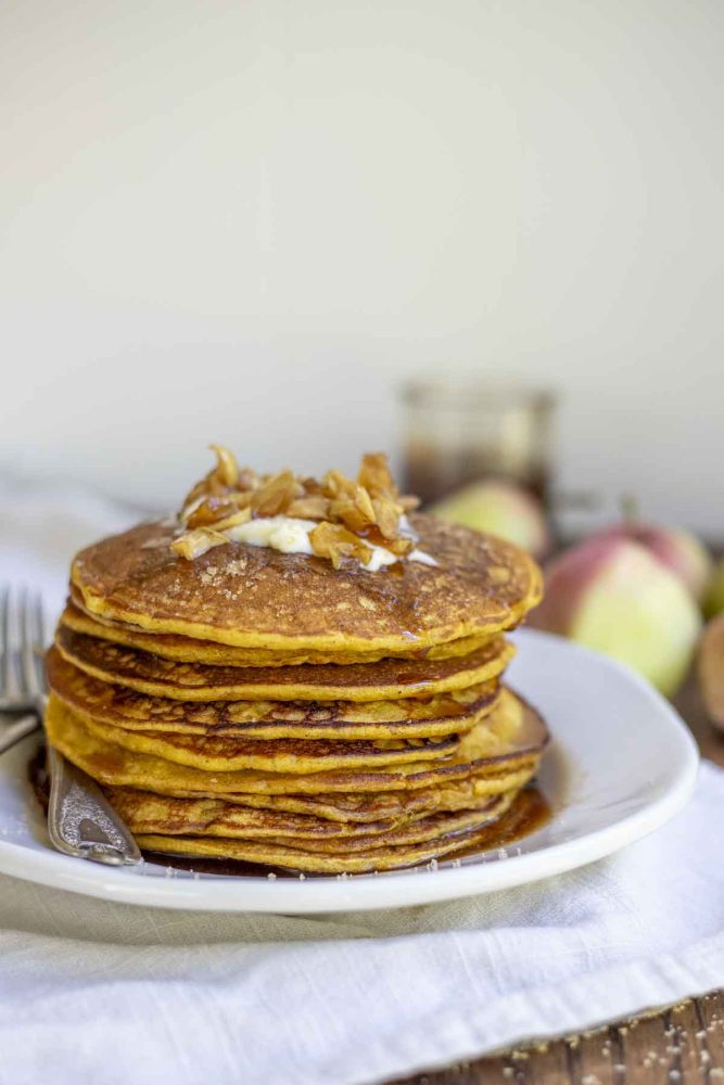 close up picture of a stack of sourdough pumpkin pancakes topped with butter, apples, and syrup on a white plate on a tan napkin.