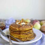 a stack of sourdough pumpkin pie pancakes topped with butter and apples on a white plate. The plate is on a white and blue stripped towel
