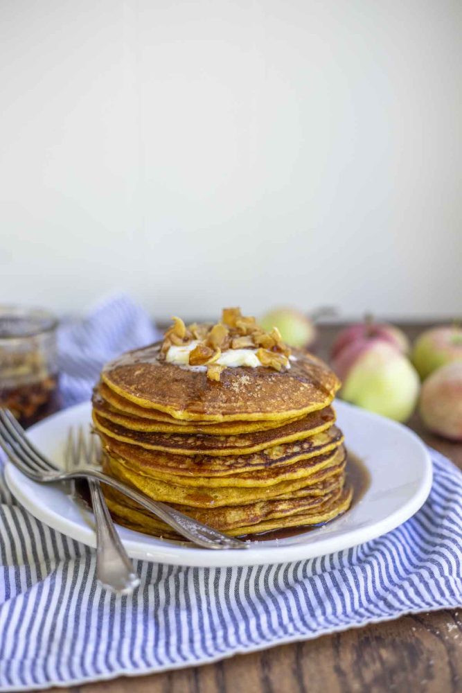 a stack of sourdough pumpkin pie pancakes topped with butter and apples on a white plate. The plate is on a white and blue stripped towel