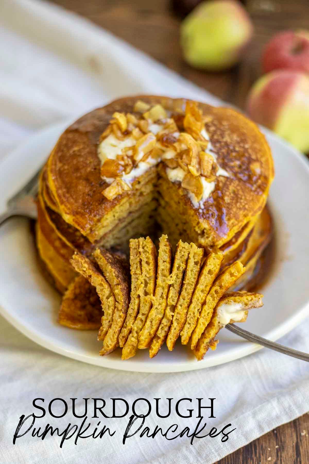 stack of sourdough pancakes topped with apples and syrup with a large slice of pancakes taken out and on a fork resting in front of the stack.