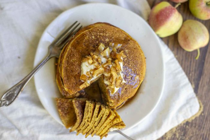 overhead photo of a stack of pumpkin sourdough pancakes topped with butter and apple syrup on a white plate. A slice taken out of the pancakes is on a fork in the front part of the plate. Apples are in the background