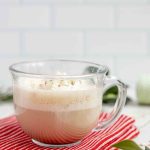 side view of a eggnog latte topped with whipped cream and sprinkles in a glass mug on a red towel on a marble countertop surrounded by sprinkles and greenery