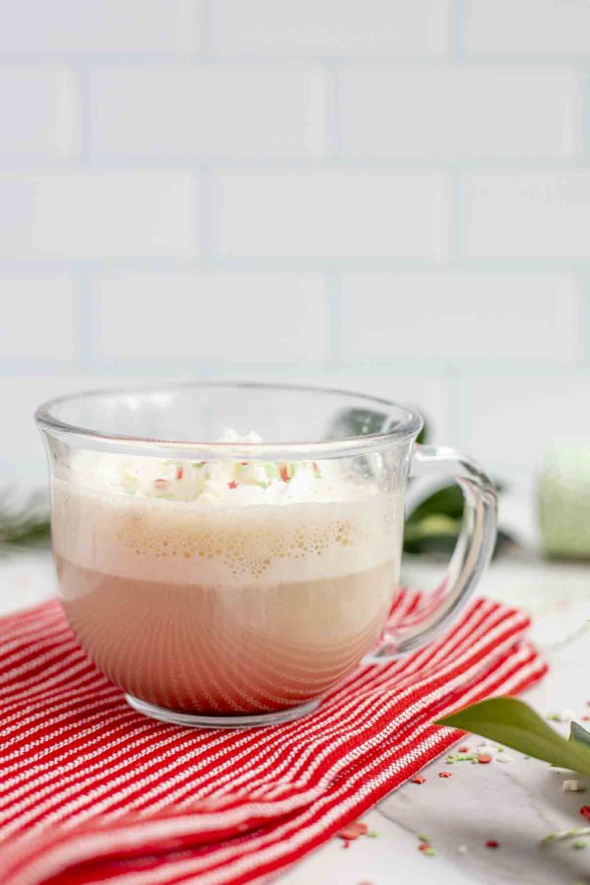 side view of a eggnog latte topped with whipped cream and sprinkles in a glass mug on a red towel on a marble countertop surrounded by sprinkles and greenery
