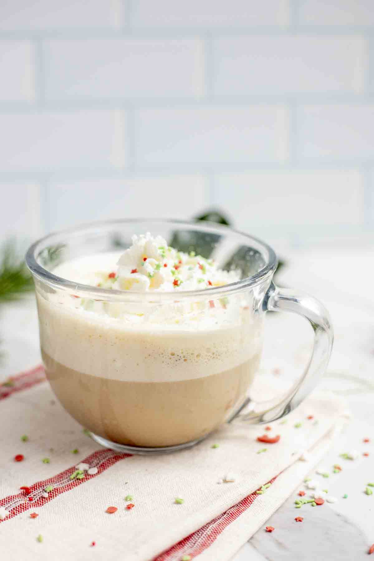 side view of a glass mug full of a eggnog latte topped with whipped cream and Christmas sprinkles. The mug sits on a white and red stripped towel with sprinkles scattered around.