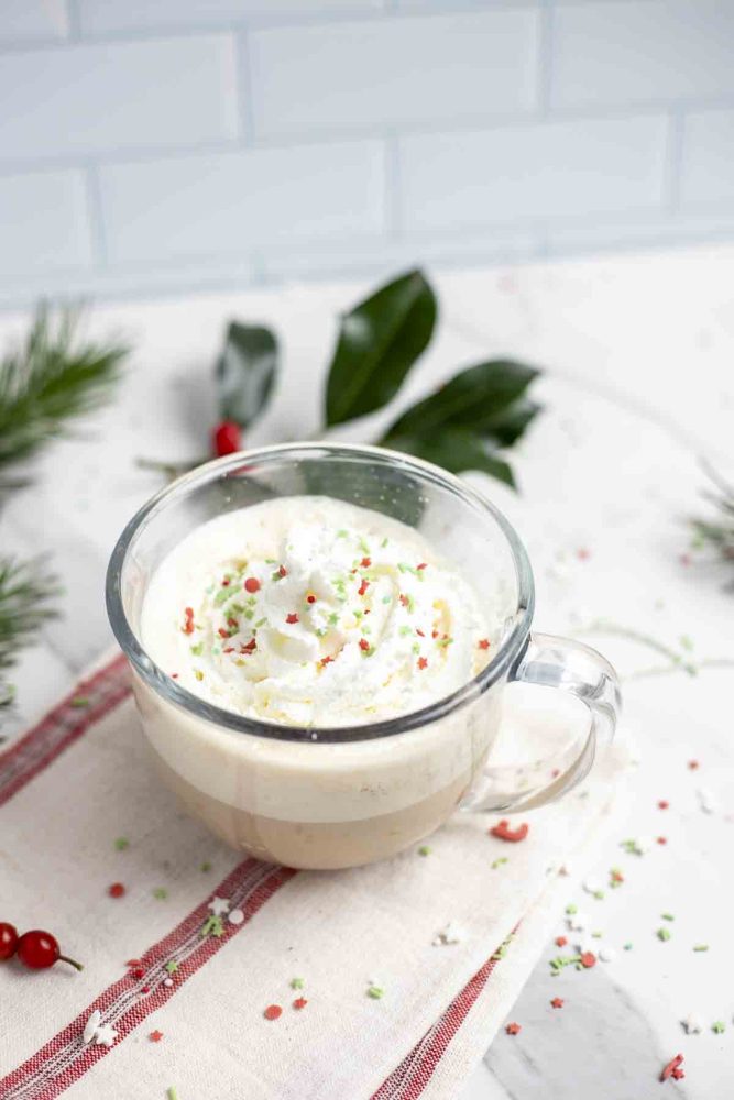 eggnog latte topped with whipped cream and sprinkles in a glass mug on top of a cream and red stripped towel surrounded by sprinkles and greenery