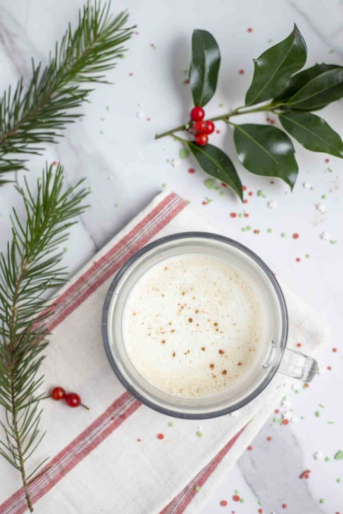 overhead photo of a eggnog latte with nutmeg sprinkled on top in a glass mug on top of a cream and red stripped towel . Greenery and holly surround the mug