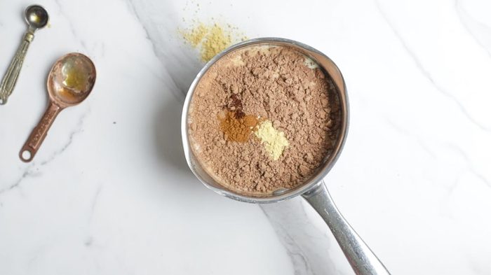 milk, spices, cocoa powder, and maple syrup in a small saucepan on a marble counter with measuring spoons in the top left corner
