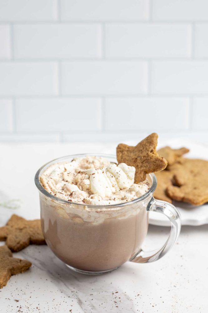 Glass mug with gingerbread hot chocolate topped with homemade whipped cream and a star shaped gingerbread cookie. The mug is on a marble counter and has gingerbread cookies to the left and a plate of cookies in the background.