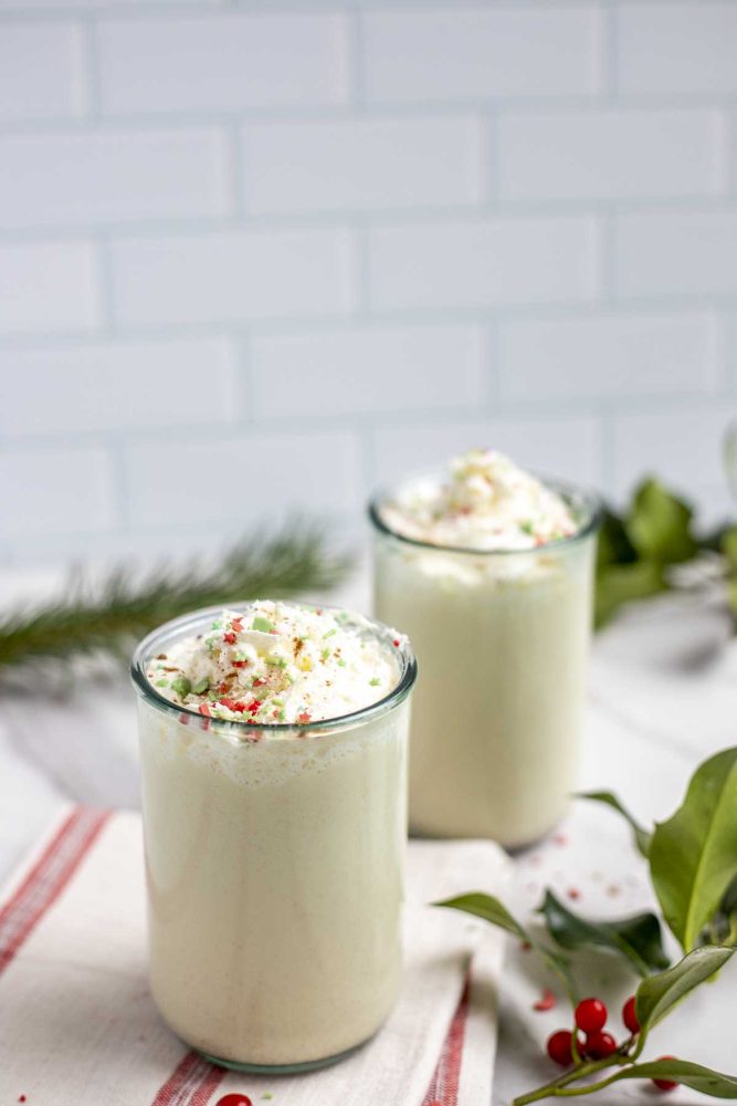 two glasses of easy homemade eggnog on a cream and red stripped towel with holly and greenery surrounding the glasses