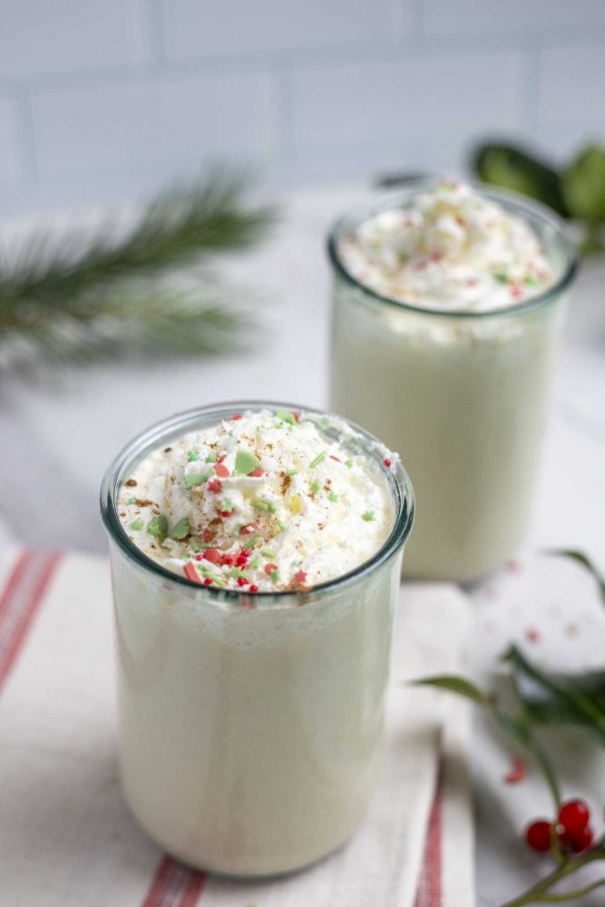 two glasses of homemade eggnog topped with whipped cream and sprinkles. The glasses are on a white and cream strolled towel and surrounded by greenery