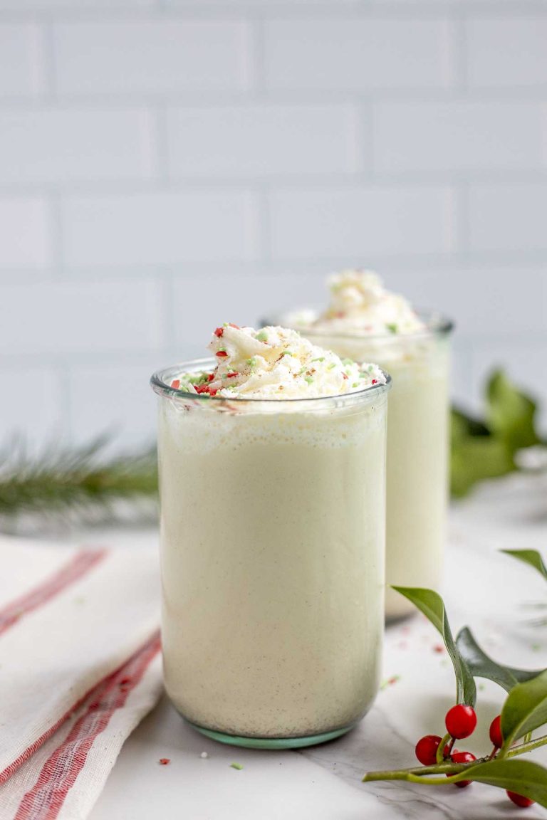 two glasses of homemade eggnog topped with whipped cream and sprinkled with christmas sprinkles on a marble countertop surrounded by greenery and a red and cream towel