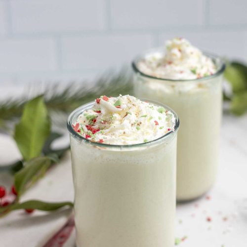 two jars of homemade eggnog topped with whipped cream and red, green, and white sprinkles on a marble countertop with greenery in the background