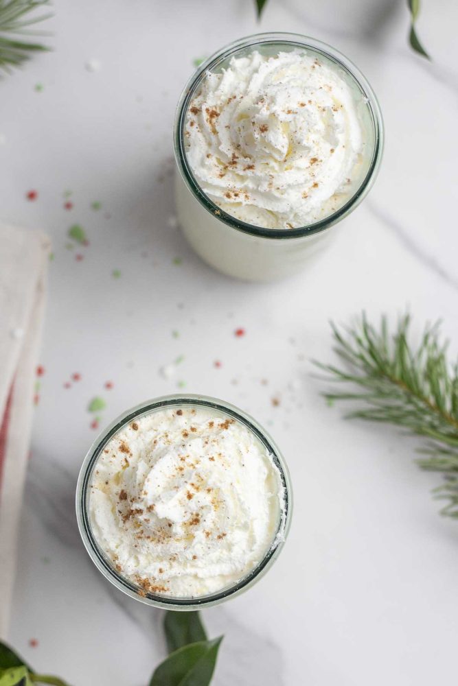 overhead photo of two glasses of eggnog on a white marble countertop surrounded by sprinkles and greenery