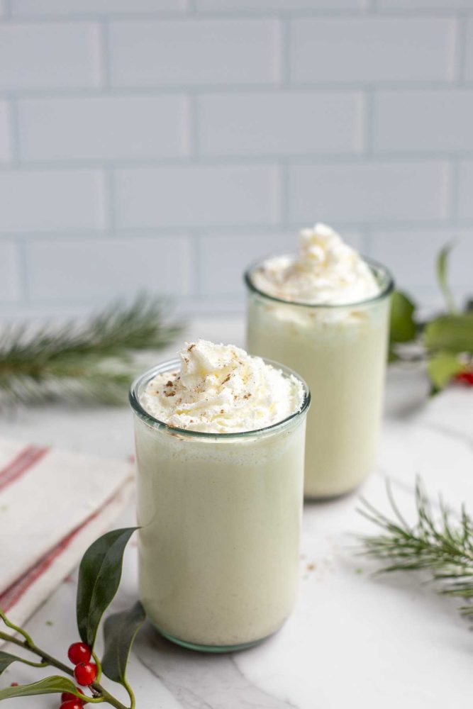two large glasses of homemade eggnog with whipped cream on marble countertop with a red and white stripped towel to the right and greenery surrounding the glasses
