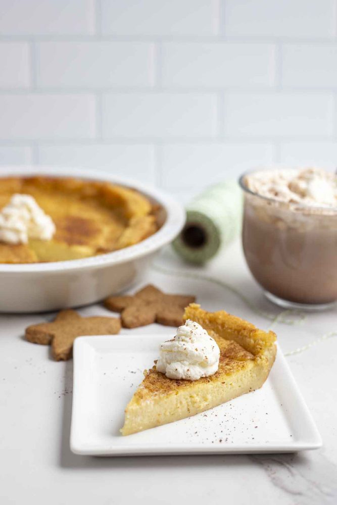 slice of custard pie with a dollop of whipped cream on a white square plate on a marble countertop. The pie plate with pie, hot chocolate, and cookies are in the background