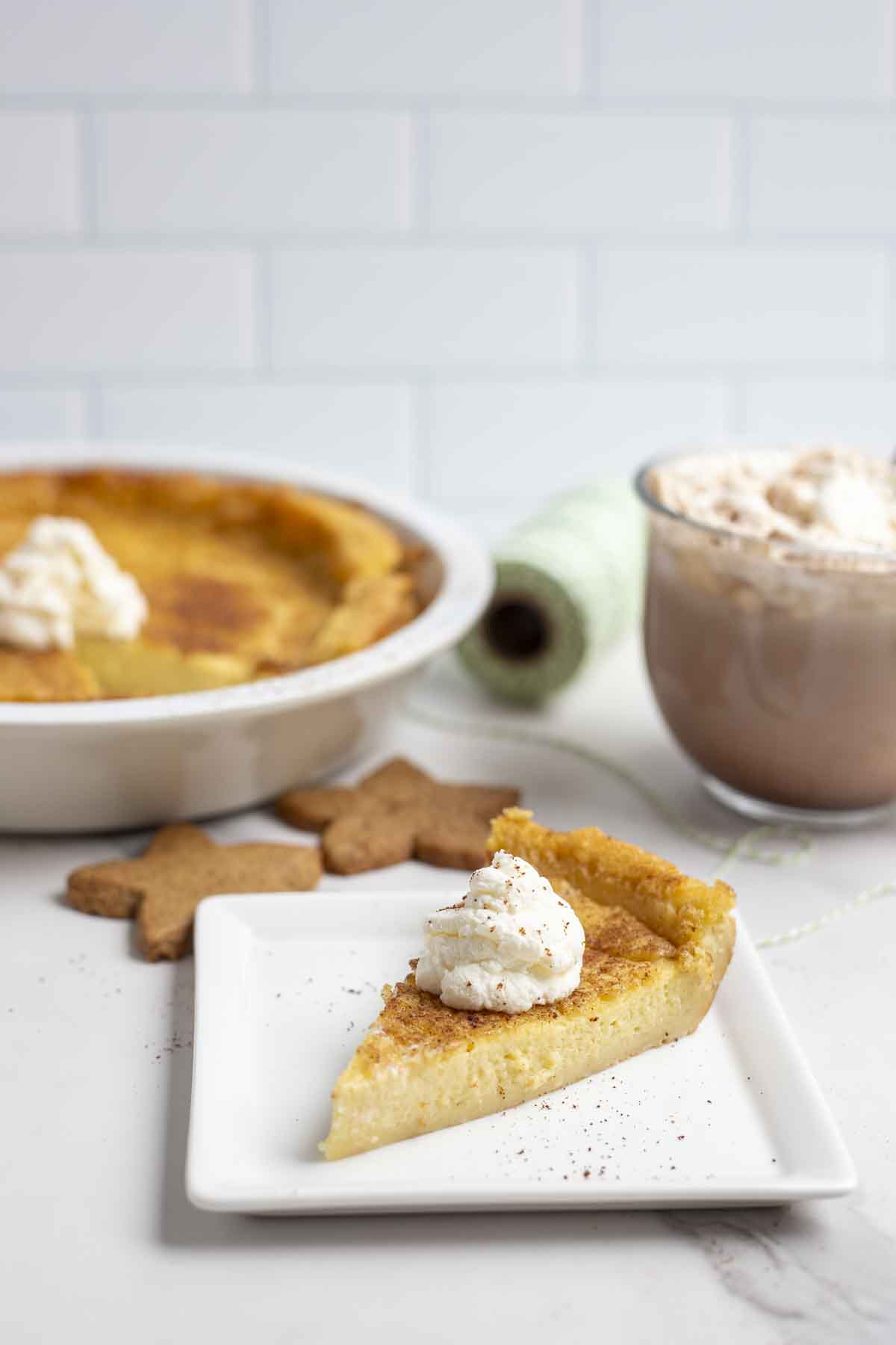 slice of custard pie with a dollop of whipped cream on a white square plate on a marble countertop. The pie plate with pie, hot chocolate, and cookies are in the background.