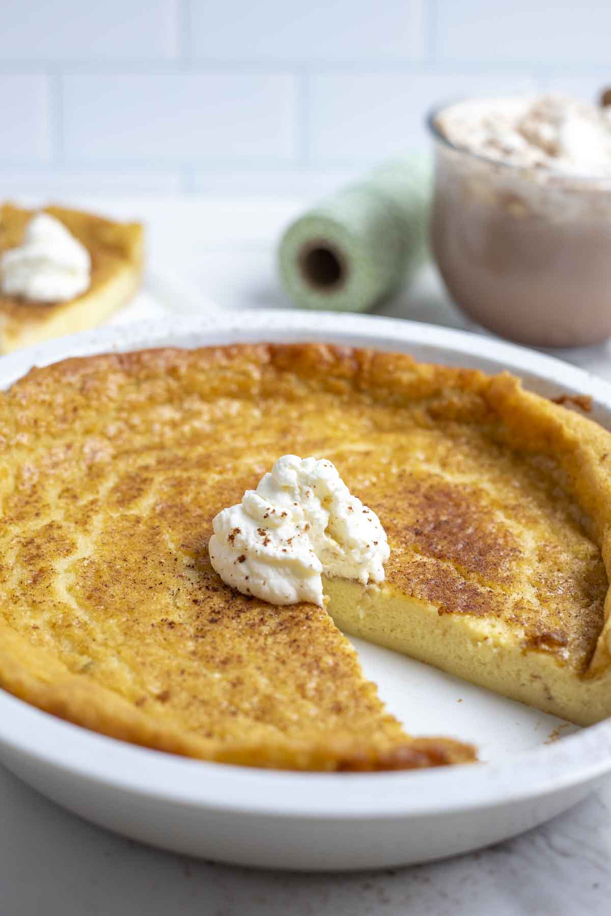 close up picture of a custard pie with a dollop of whipped cream and a slice taken out. Mug of hot chocolate, spool of green string, and a piece pie is in the background.