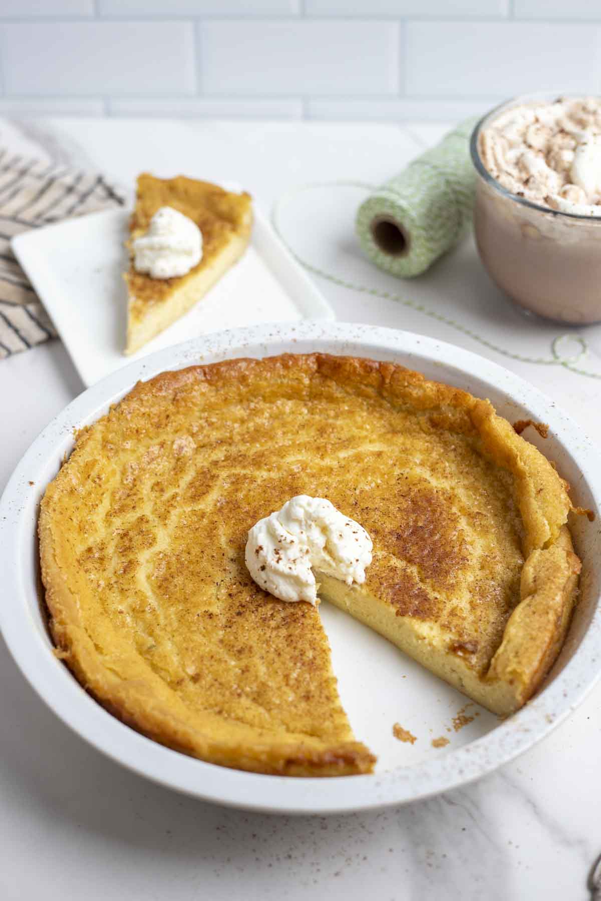 custard pie with a slice taken out and a dollop of whipped cream in a white pie plate. The pie is on a marble counter with a slice of pie, hot chocolate, and green string in the background.