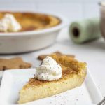 slice of custard pie topped with whipped cream on a white plate with a pie plate with more pie in the background