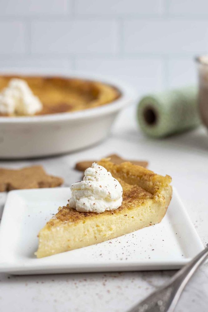 slice of custard pie topped with whipped cream on a white plate with a pie plate with more pie in the background