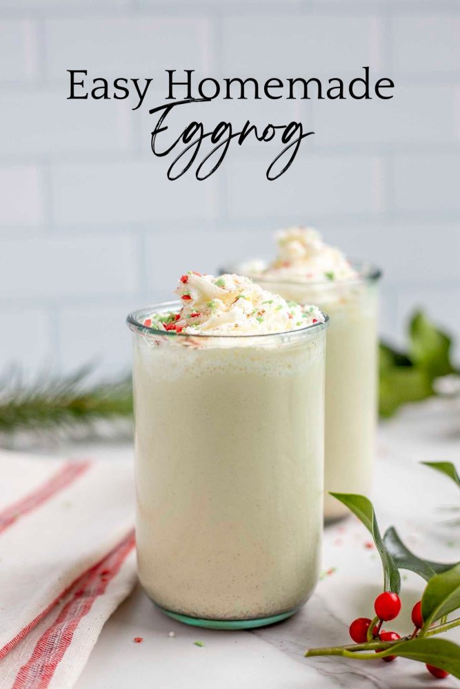 two glasses of homemade eggnog topped with whipped cream and sprinkled with christmas sprinkles on a marble countertop surrounded by greenery and a red and cream towel