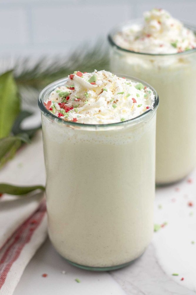two glasses of eggnog topped with whipped cream and sprinkles on a white marble countertop