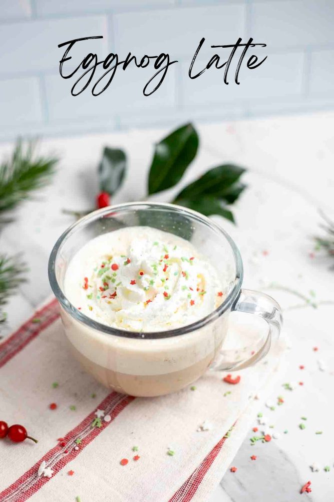 eggnog latte topped with whipped cream and sprinkles in a glass mug on top of a cream and red stripped towel surrounded by sprinkles and greenery