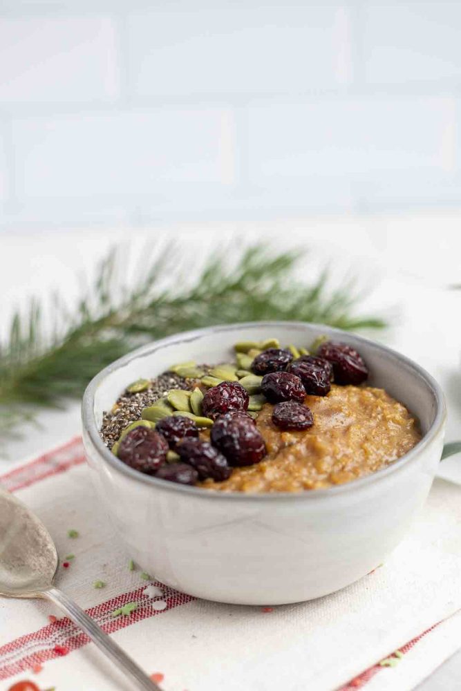 side view of gingerbread oatmeal topped with seeds and dried cranberries in a stoneware bowl on a white and cream stripped towel. Fresh greenery is in the background and vintage spoon to the left