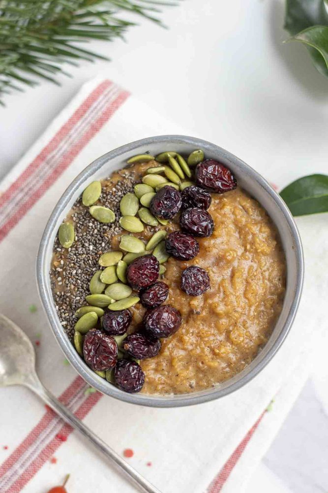 overhead photo of a bowl of gingerbread oatmeal topped with pumpkin seeds, chia seeds, and raisins on a white and cream stripped towel with greenery in the background