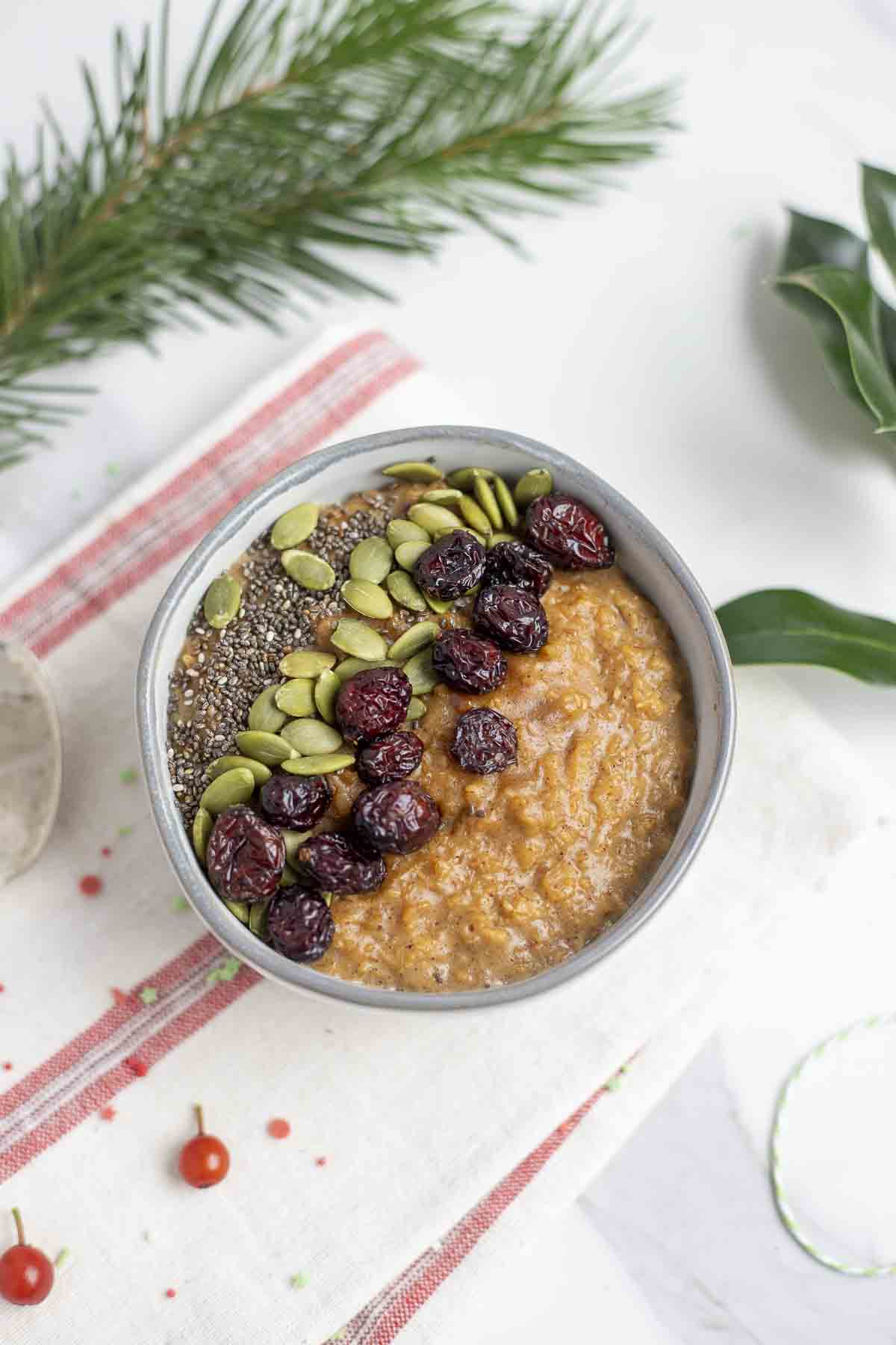 overhead photo of a bowl of gingerbread oatmeal topped with chia seeds, pepitas, and raisins on a red and cream stripped towel and surrounded by fresh greenery.