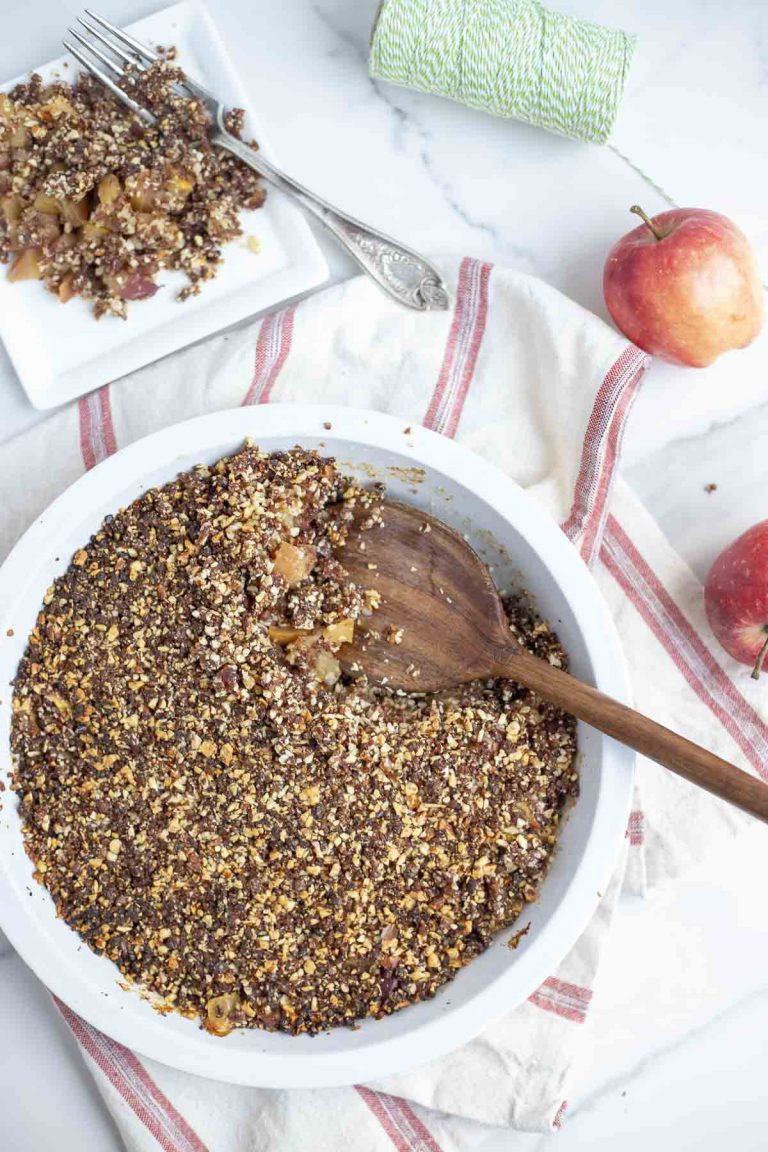 overhead photo of a healthy apple crisp in a pie plate with a wooden spoon in the dish. The pie dish is on a white and red stripped towel with a plate of crisp and apples in the background