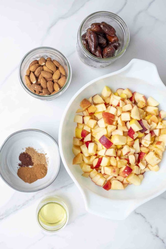 bowls and jars filled with apples, dates, almonds, spices and oil on a marble countertop