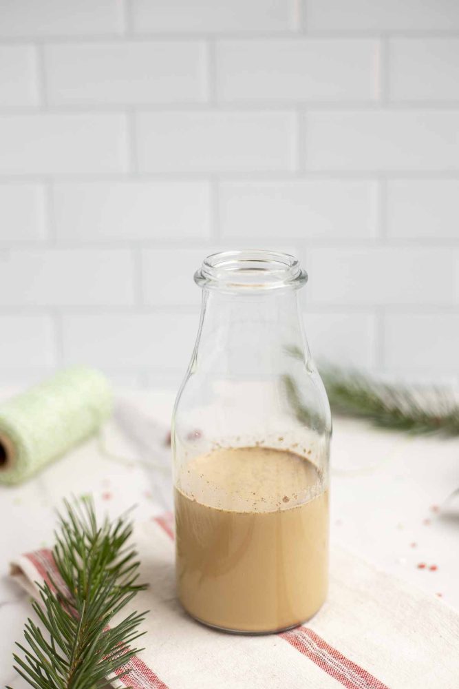 a small milk jar of gingerbread creamer on a white and red stripped towel with greenery to the left and a spoon of green thread in the background
