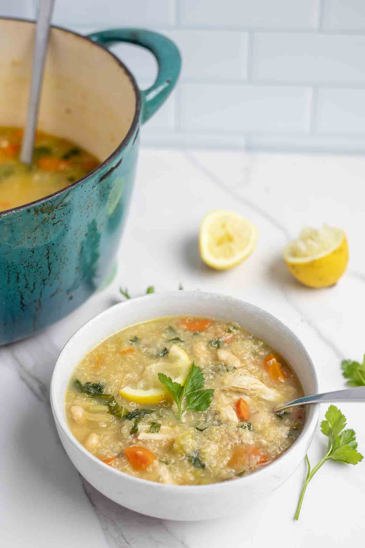 Bowl of lemon chicken quinoa soup topped with a slice of lemon and fresh parsley in a white bowl on a white countertop. a whole lemon sliced in half and a turquoise pot in the background