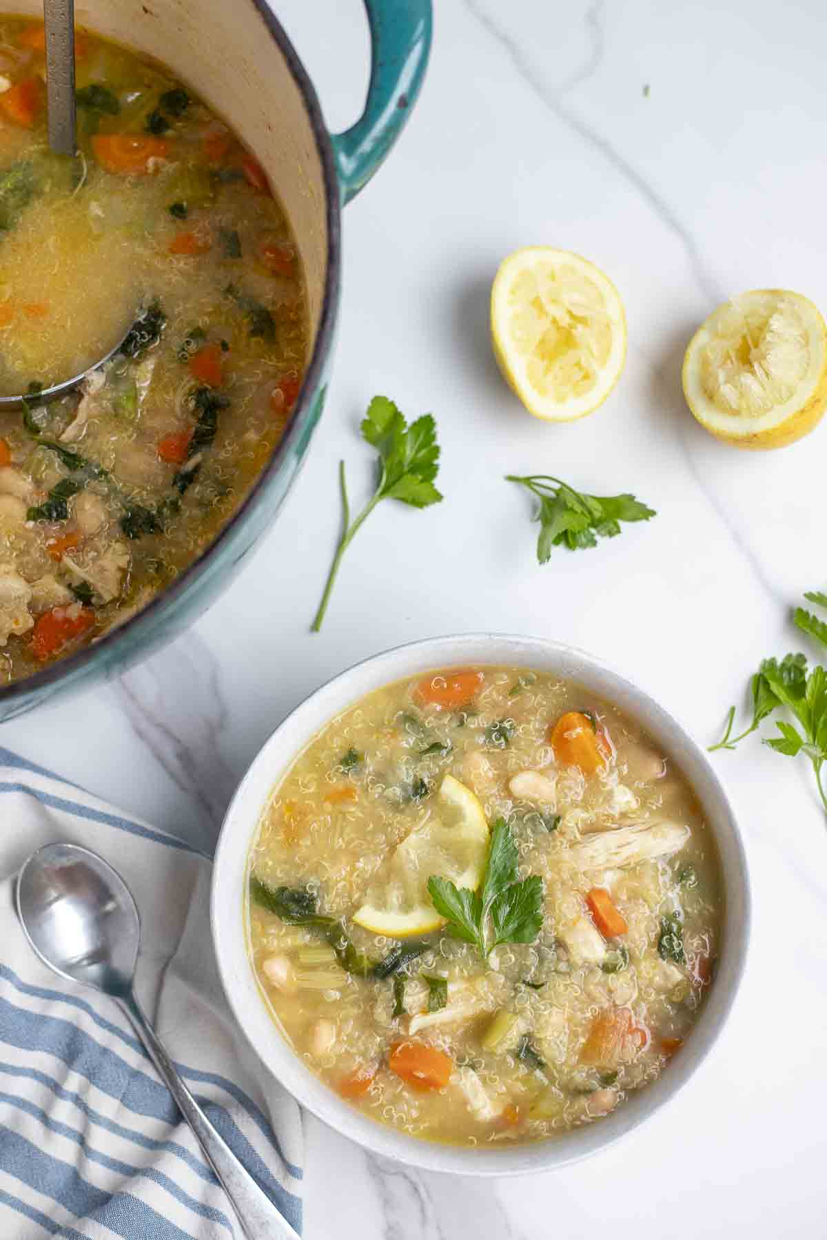 overhead photo of a bowl of chicken quinoa soup on a white countertop surrounded by two lemon halves, fresh parsley, a spoon, blue and cream stripped napkin and a dutch oven filled with more soup.