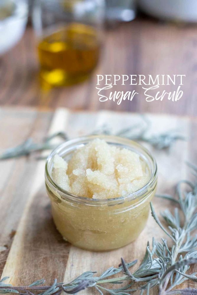 Homemade peppermint sugar scrub in a glass mason jar on a wood cutting board surrounded by lavender. A jar of oil is in the background