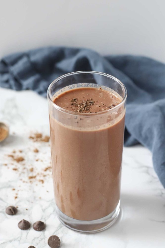 tall glass of chocolate cauliflower smoothie topped with chopped chocolate. The glass is on a white marble countertop and surrounded by chocolate chips, cocoa powder, and a blue napkin in the background