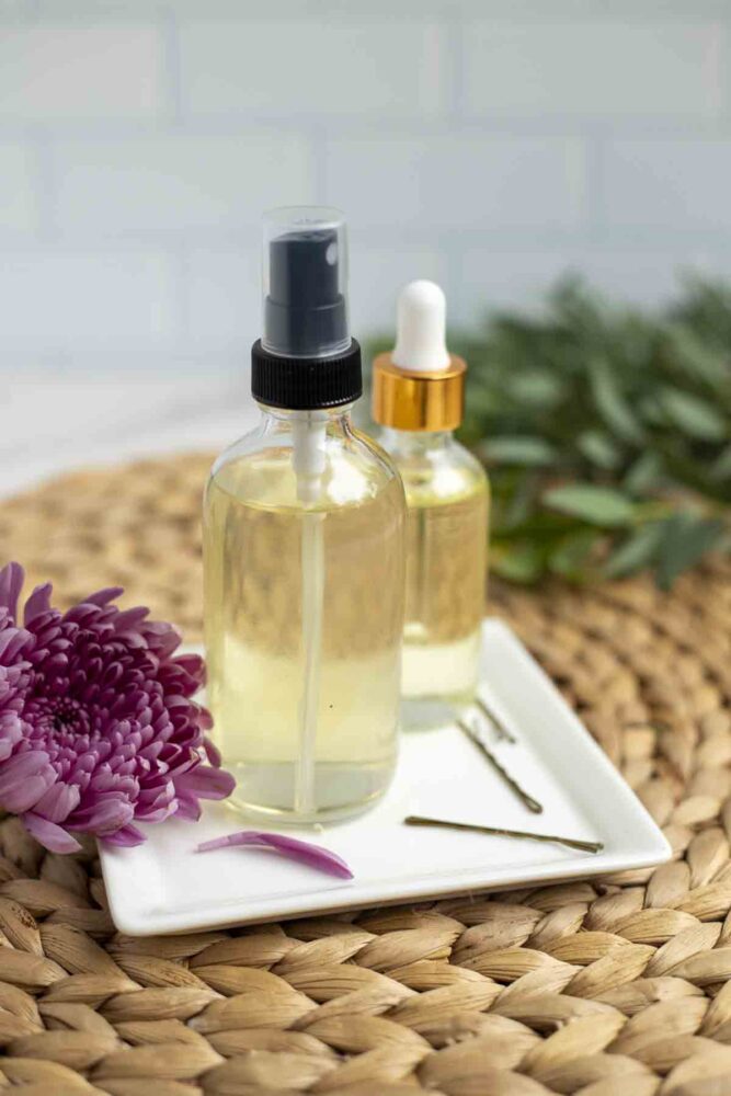 homemade hair spray and hair serum on a white plate with bobby-pins on a woven mat. A pink flower and greenery surround the bottles