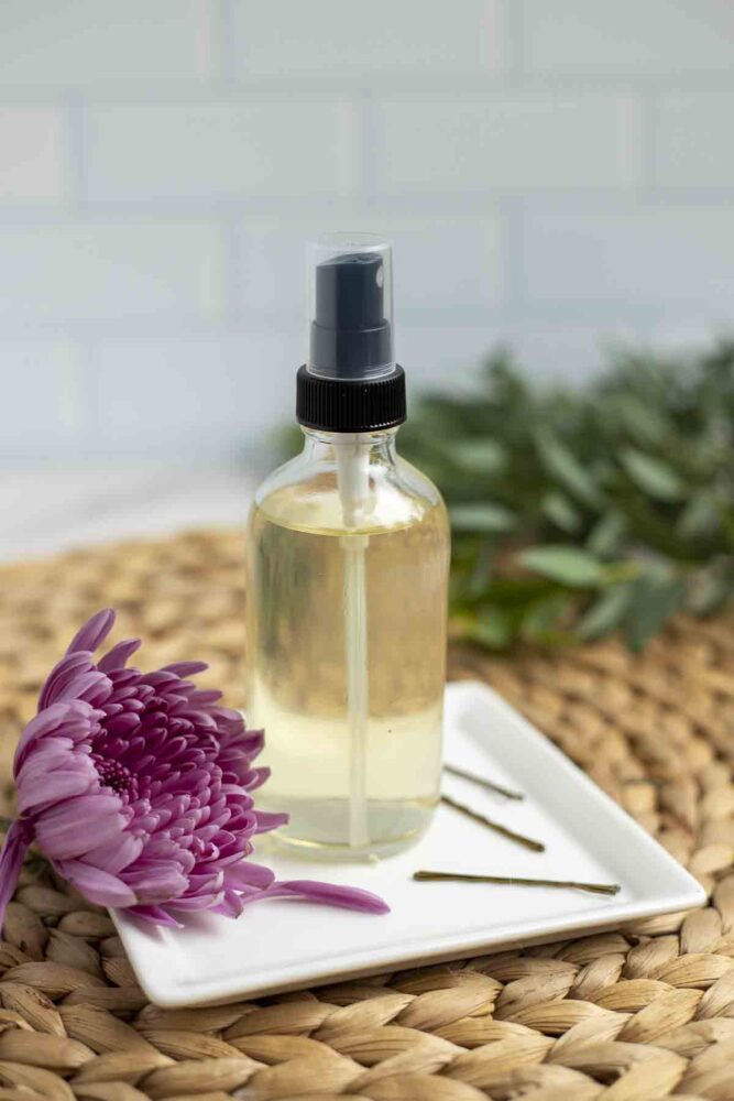 homemade hairspray in a glass bottle on a white plate with a pink flower to the right and bobby pins to the left. The plate is on a woven placemat with greenery in the background